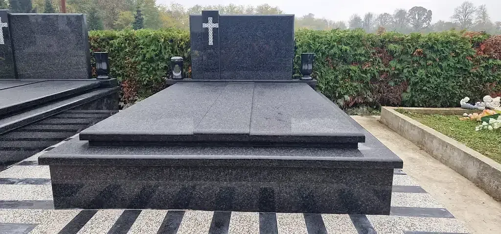 Crni granitni grob s ugraviranim križem i dvama lampionima ispred zelene živice