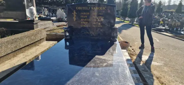Tamni polirani grob s valovitom nadgrobnom pločom uz grobljansku stazu