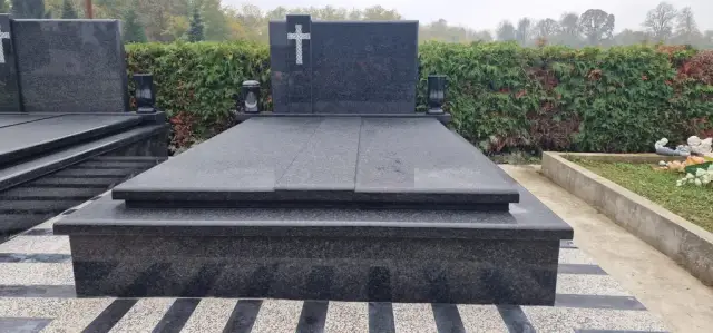 Tamnosivi dvojni grob s bijelim križem ispred zelene živice